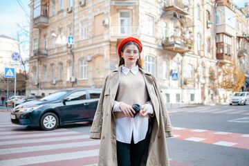 Happy young caucasian woman in good mood walks with coffee around city during day. Brunette wears glasses and red beret. Real emotions concept.  