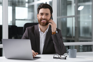 Man talking on phone while working with laptop at white desk in office