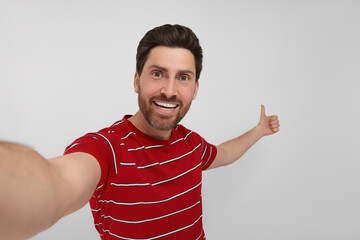 Fototapeta premium Smiling man taking selfie and showing thumbs up on white background
