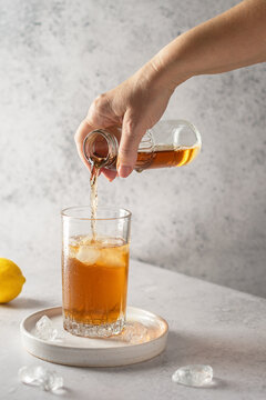 A woman's hand pours cold tea from a small bottle into a glass with ice. On a gray stone table are pieces of ice and a lemon. Refreshing drink in summer.