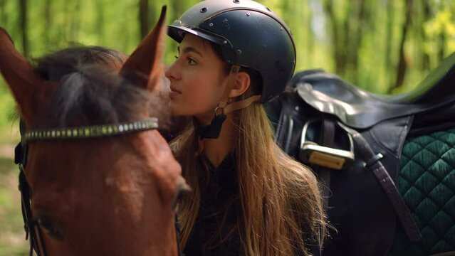 Slim pretty woman kissing neck of brown horse talking to pet in slow motion. Portrait of cute confident Caucasian female equestrian with graceful purebred stallion in sunny forest outdoors