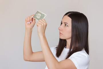 young dark-haired Caucasian woman with a serious face in a white T-shirt holding a dollar bill in her hands and checking it for authenticity. The concept of financial literacy