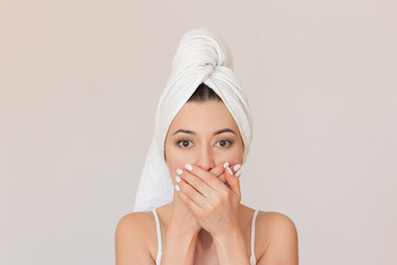 close-up photo of a young pretty dark-haired woman with a white towel wrapped around her head, who covering her mouth with both hands in surprise. Isolated on a beige background.