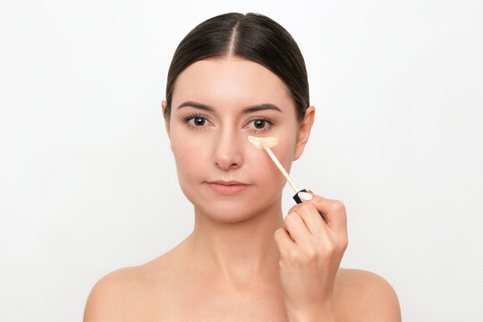 A young attractive Caucasian dark-haired woman applying a concealer under her eyes from dark circles under her eyes. Isolated on a light background