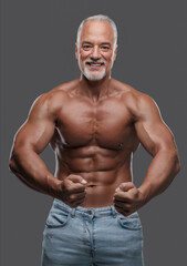 Fototapeta na wymiar Handsome older man with a stylish grey beard, muscular physique, ripped jeans and bare torso, posing against a grey backdrop
