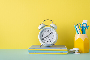 Back to school concept. Photo of school accessories on blue desk alarm clock over stack of copybooks mini stapler and stand for pens on yellow wall background