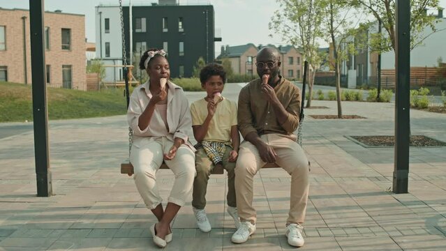 Stab shot of delighted African American family of three talking and eating ice cream while sitting on wooden playground swing outside in summer
