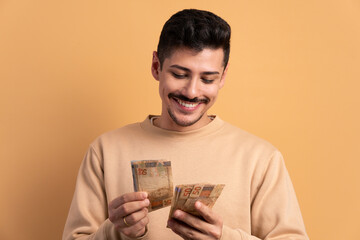 happy brazilian man looking at brazilian money cash in beige studio background. financial, credit,...