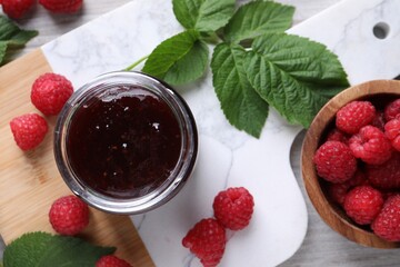 Delicious raspberry jam, fresh berries and green leaves on table, flat lay