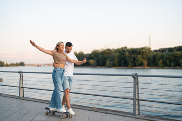 Smiling cheerful young couple with skateboard. He is teaching her to drive longboard she is happy and letting go her hand. Young woman with hands in the air enjoying skateboard ride with her boyfriend
