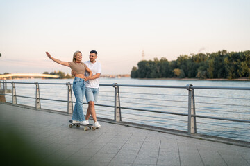 Couple being happy on skateboard. She let go one hand and he is holding her waist. Both are widely smiling.