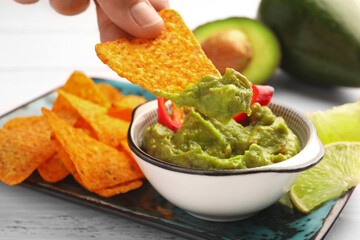 Woman dipping delicious nachos chip into guacamole at white wooden table, closeup