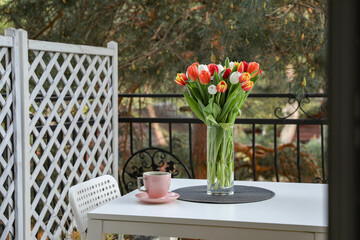 Beautiful colorful tulips in glass vase and cup of drink on white table at balcony