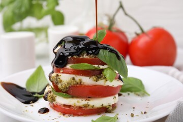Pouring balsamic vinegar onto stacked Caprese salad with pesto sauce on table, closeup