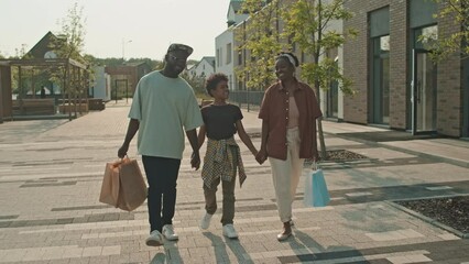 Full length shot of joyful African American family of three carrying paper shopping bags talking while walking along street after shopping together on sunny day
