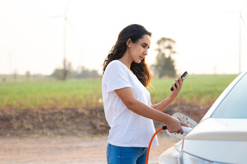 World Environment Day. save world save life. Hispanic female driver electric car holding electrical plug connector to charge the batteries hybrid car at home or charging station. Using smartphone