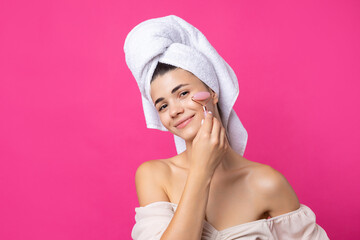 A beautiful cheerful attractive girl with a towel on her head holds a cosmetic roller near her face against a pink background.