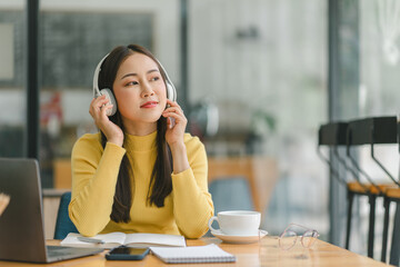 Focused Asian businesswoman wearing headphones is taking notes in a notebook while watching a webinar video course. Serious female student listening to the lecture to study online through e-learning.