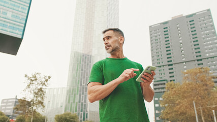 Mature male tourist wearing casual clothes, standing on modern buildings background, looking for right direction on smartphone, using map app