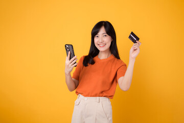 Independent Asian woman 30s in orange shirt, using smartphone and holding credit card on yellow background. Flexible online payment.