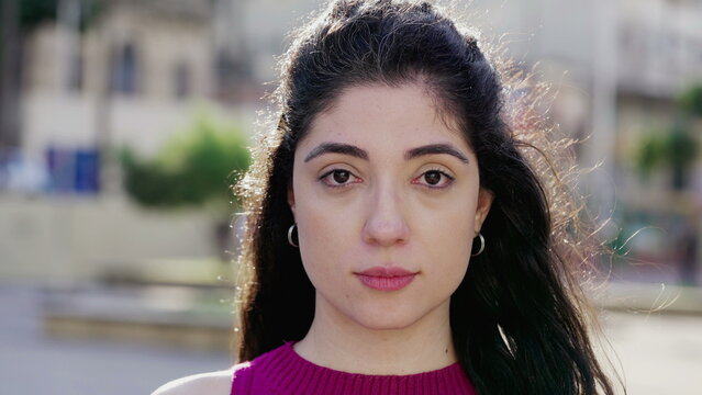 One young woman with neutral expression looking at camera standing outside in city park. A female millennial 30s person close-up face