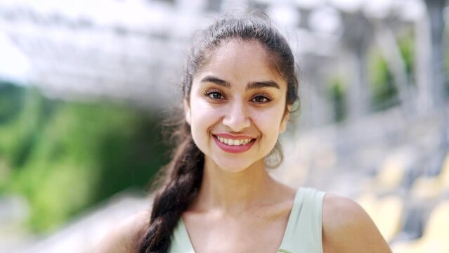 Portrait of a young smiling sporty fit woman at urban city stadium. Happy fitness female girl atlet standing outdoors looking at camera. Head shot of positive motivated female trainer or coach Closeup
