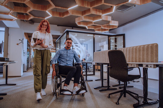 Young business colleagues, collaborative business colleagues, including a person in a wheelchair, walk past a modern glass office corridor, illustrating diversity, teamwork and empowerment in the