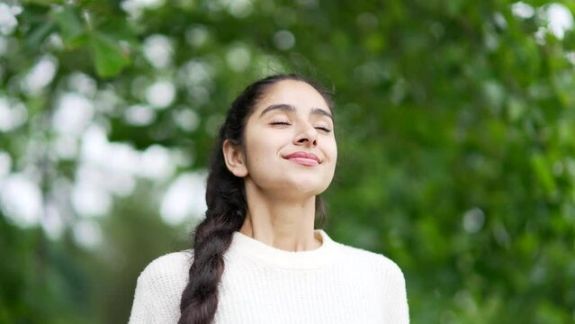 Young indian woman enjoying nature taking deep breaths of fresh air standing among trees in forest or park. Outdoors closing Female girl Inhaling and Exhaling her eyes calm rest and stress relief