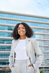 Happy young African American business woman wearing suit and glasses looking at camera standing in city. Smiling ethnic female sales professional, executive manager, office employee vertical portrait.