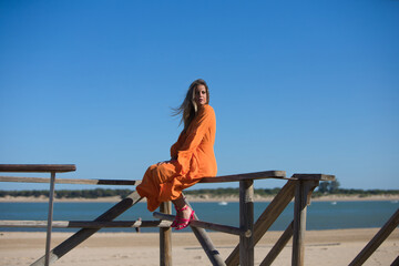 Pretty young blonde woman in orange dress sitting on the wooden railing of the beach promenade. In the background on the horizon blue sky and sea. Holiday and travel concept.