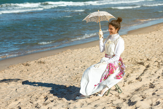young woman sitting on the shore of the beach dressed in period costume with parasol and hat