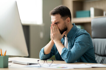 Work Burnout. Tired Young Businessman Sitting At Desk In Office, Rubbing Eyes
