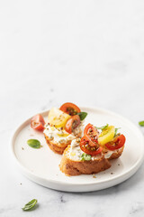 Bruschetta sandwiches with tomatoes, cream cheese, olive oil and basil on a plate on white marble background, text space. Traditional italian antipasti