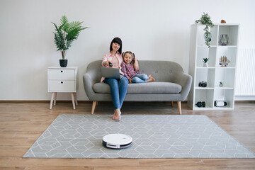 Attractive woman programing robotic vacuum via remote control while sitting behind laptop with kid on sofa. Caucasian mother and daughter watching cartoons online while tech gadget cleaning carpet.