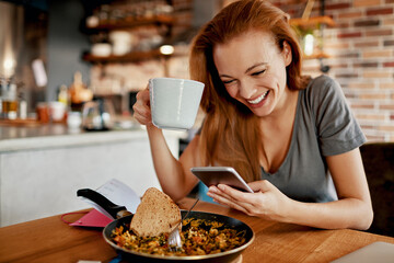 Young woman using a smart phone while having a healthy breakfast in the morning in her kitchen