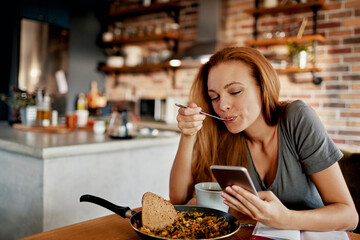 Young woman using a smart phone while having a healthy breakfast in the morning in her kitchen