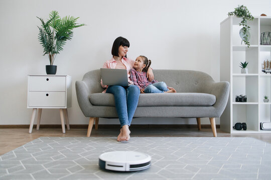 Pretty mother and daughter resting on sofa with computer while robot cleaner handling debris on floor. Happy caucasian ladies scrolling digital photo album on portable gadget with smart home device.