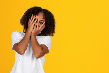 Shocked scared shy teenage black girl in white t-shirt covers face with hands, isolated on yellow background