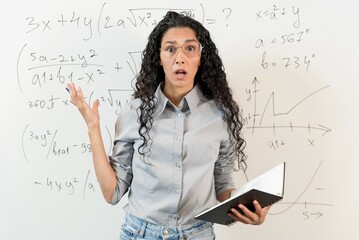 Misunderstanding, stress, fatigue, burnout. Curly haired female teacher in glasses shrug her hands, looks at the camera in surprise. Background is a blackboard with mathematical graphs and equations.