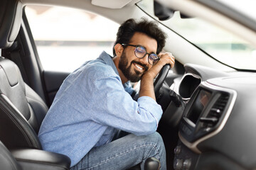 Thrilled young indian guy hugging steering wheel of new car