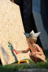 A little blond boy paints the wall with a roller.A kid in a T-shirt, shorts and a paper hat in the...