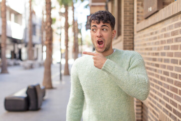 young handsome man feeling joyful and surprised, smiling with a shocked expression and pointing to the side