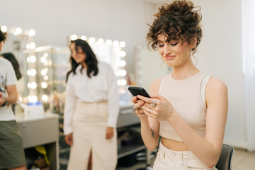 Smiling young woman with perfect makeup and hairstyle typing online message using smartphone looking to screen, on blurred background of cheerful makeup artist and hairdresser in modern beauty studio.
