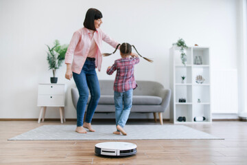 Cheerful lady in casual clothes twirling little girl around during home dancing party with working robot cleaner on floor. Happy female parent and preteen child entertaining while doing home care.