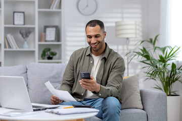 Young smiling African-American man sitting on sofa at home, holding phone and documents, working on laptop