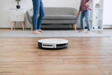 Close up view of modern cleaning robot vacuuming bare laminate floor with cropped view of females in background. Automatic gadget dusting off surface while mom and child dancing for music at home.