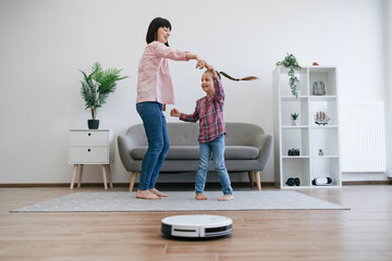 Joyful mother turning small kid around to energetic tunes while robot cleaner dealing with dirt on floor in apartment. Happy family ladies transforming household chores into engaging indoor activity.