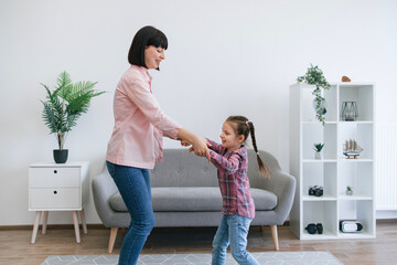 Smiling caucasian ladies in jeans outfits bouncing with rhythm of loud music while having leisure of time with each other. Cheerful mom and preteen girl releasing emotions through movements together.