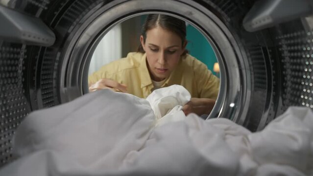 View from inside the washing machine, young woman takes out the clean white sheet from the washer