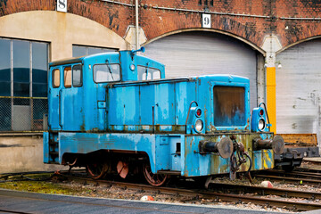 An old blue train locomotive stands on the railroad tracks next to the brick depot building